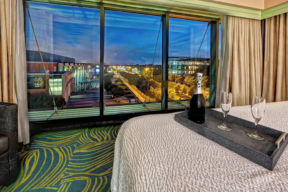 A hotel room with a large window, a bed, a bedside table with two glasses, and a patterned carpet.