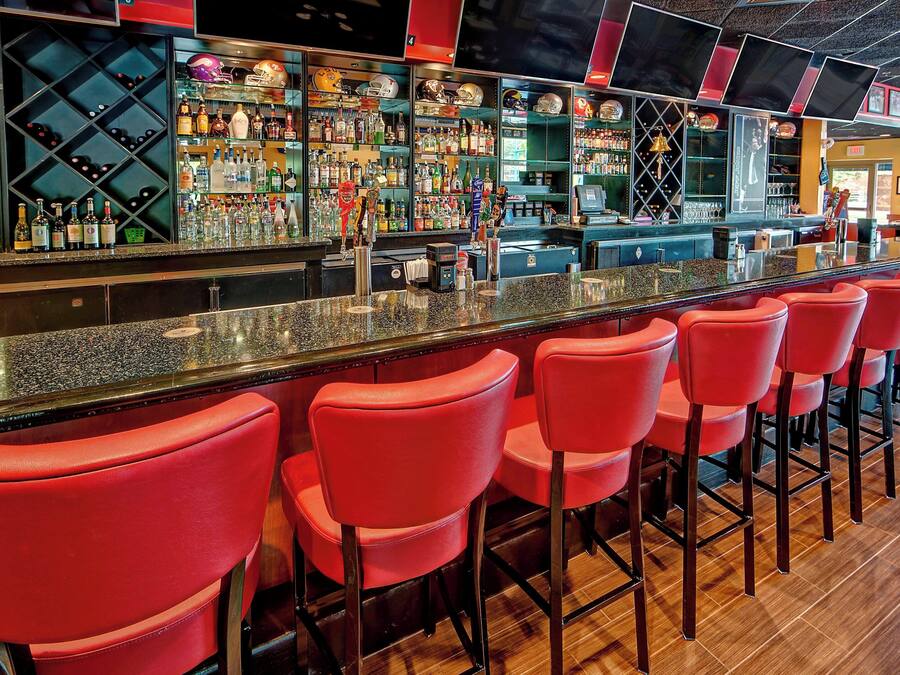 Een bar met rode barstoelen, diverse flessen en glazen op planken, en een toonbank met kassa.