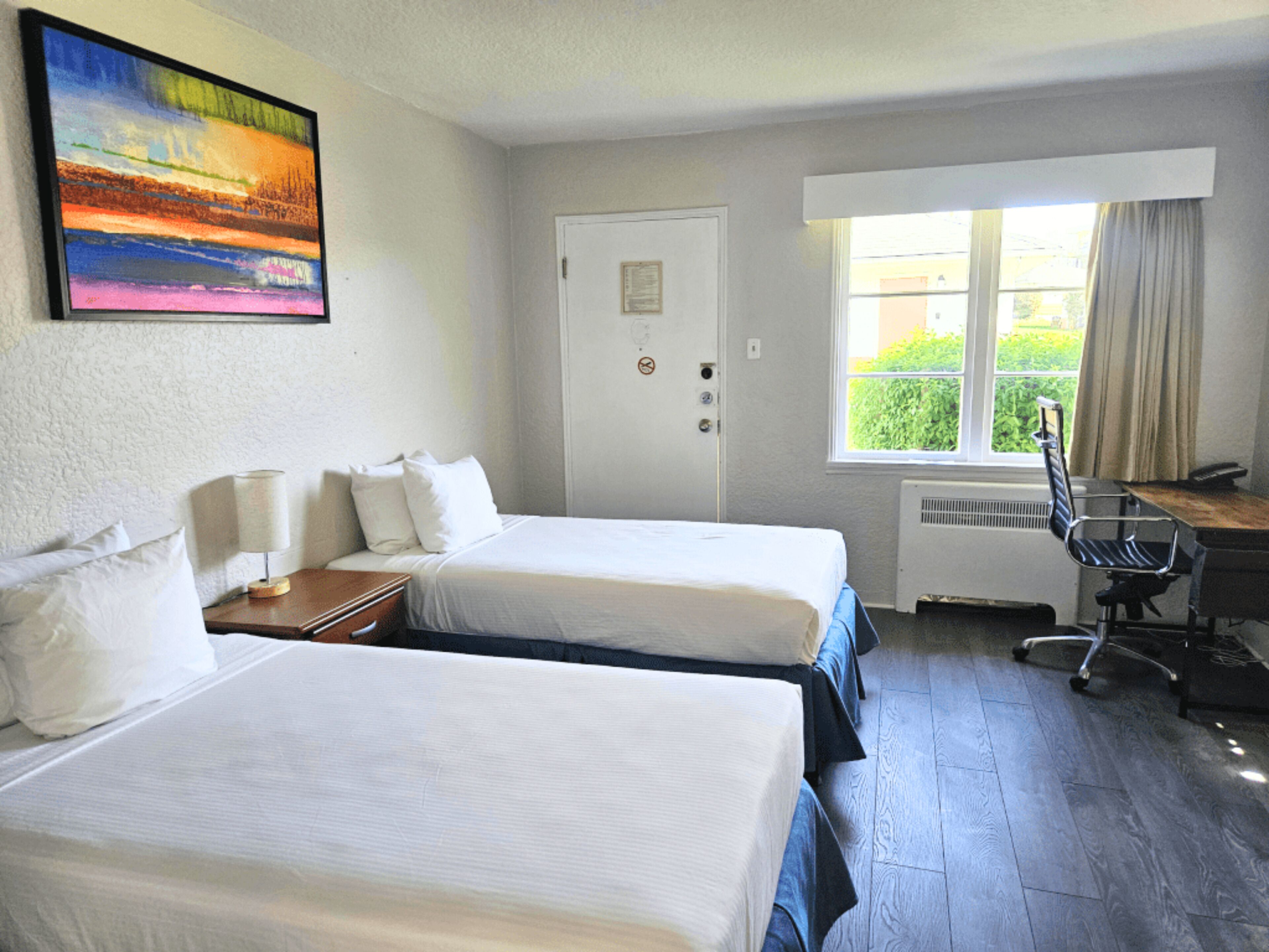 A hotel room with two beds, a desk, a chair, a radiator, and a window with a view of greenery.
