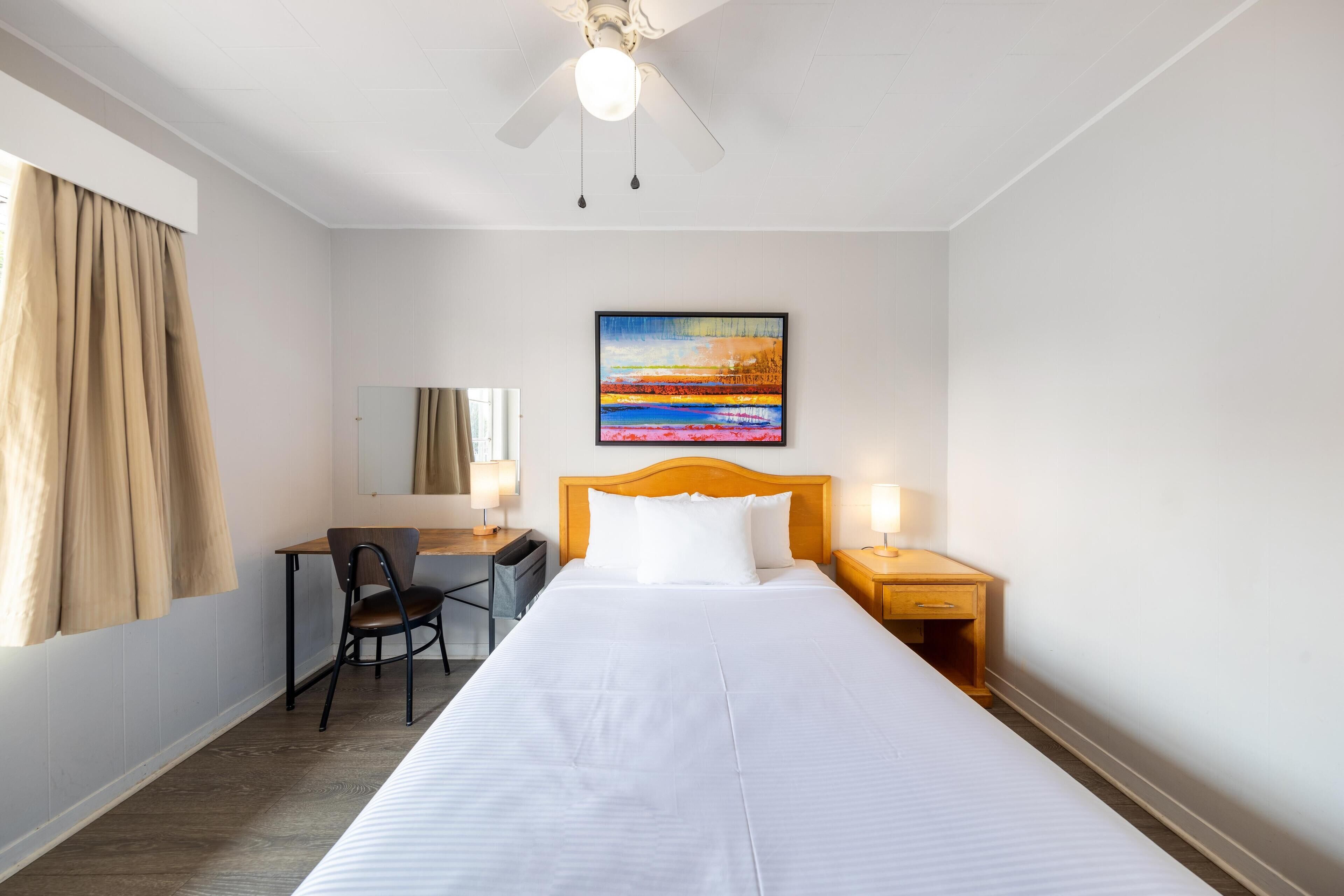 Habitación de hotel con cama, escritorio, silla, mesita de noche, ventilador de techo, un cuadro y ventana con cortinas.