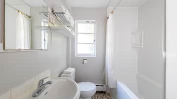 A compact bathroom with a white sink, toilet, and shower. There is a window providing natural light, and a towel rack above the sink.