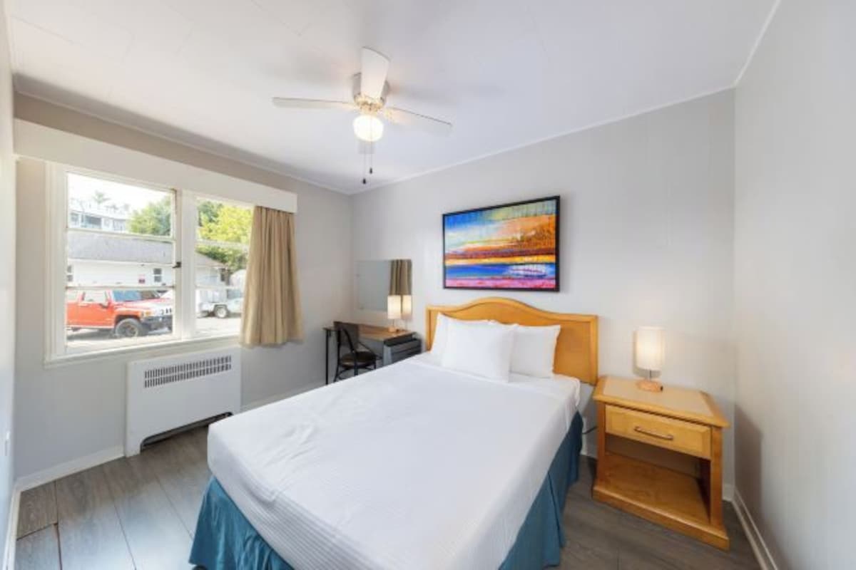 A hotel room with a bed, a desk, a chair, a nightstand, a painting, and a window with a view of the outside.