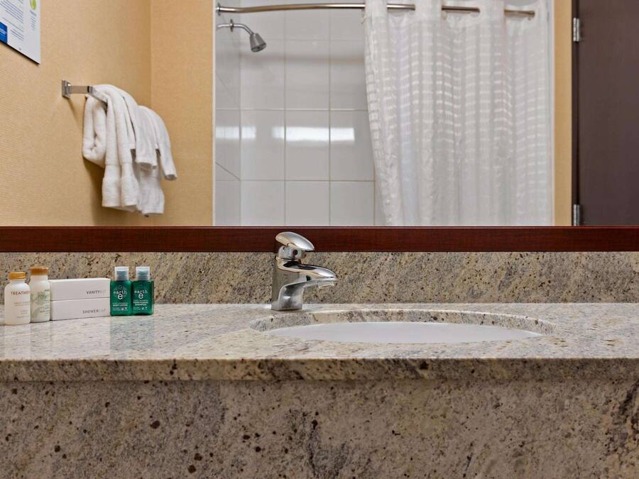 Un baño con encimera de granito, un lavabo y un espejo. Hay toallas colgadas en un perchero, y se ve una cortina de ducha al fondo.