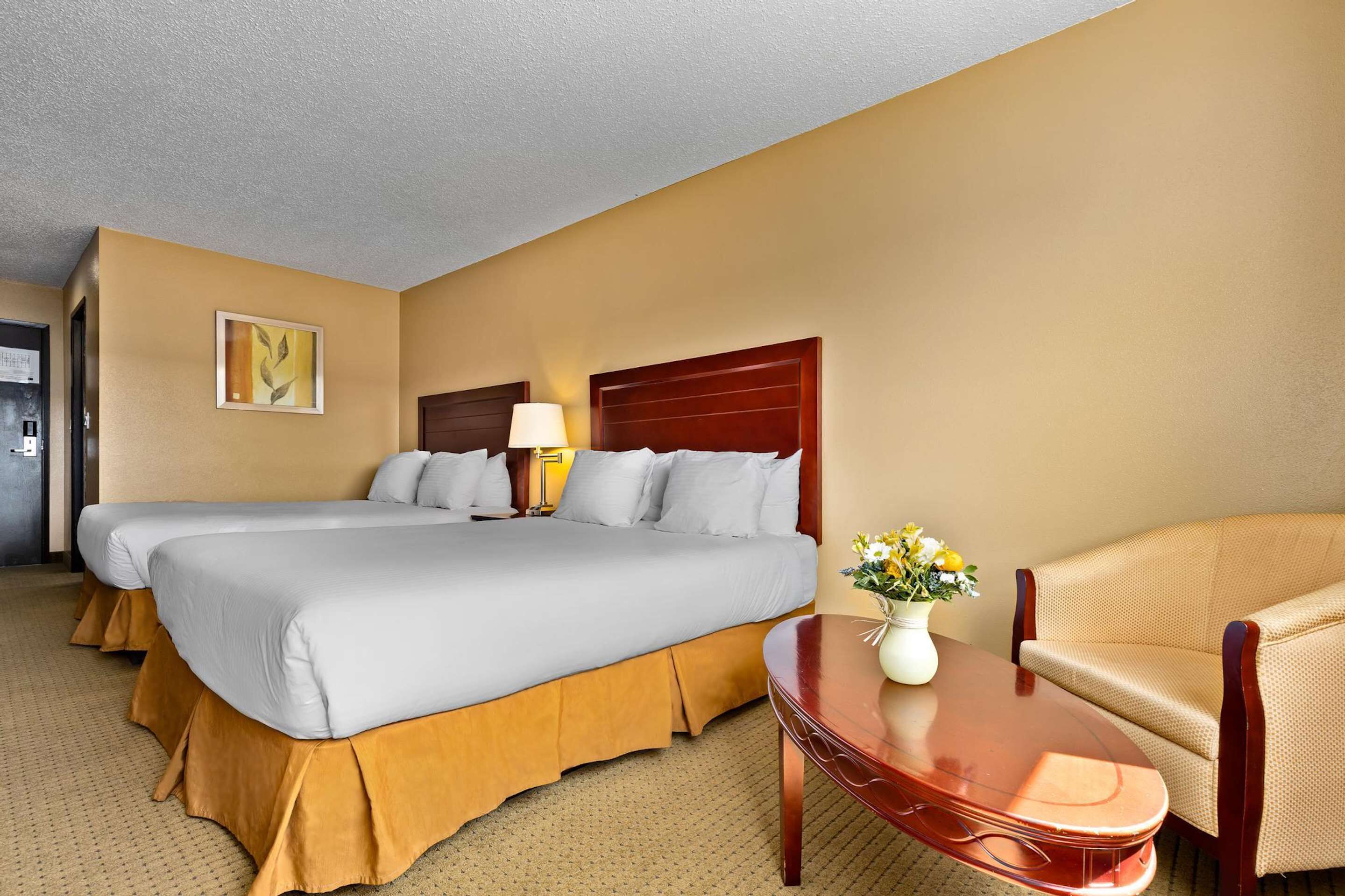 Habitación de hotel con dos camas, cabecero de madera, mesita de noche con un jarrón de flores y sofá beige.