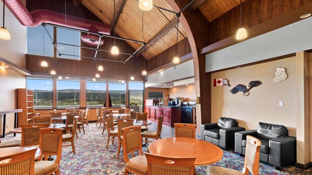 A spacious dining area with wooden tables and chairs, a patterned carpet, and large windows offering a view of the outdoors. - Coast Fraser Inn (Williams Lake)