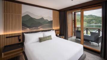 Quarto de hotel com uma cama grande, vista para um lago e varanda com cadeiras.