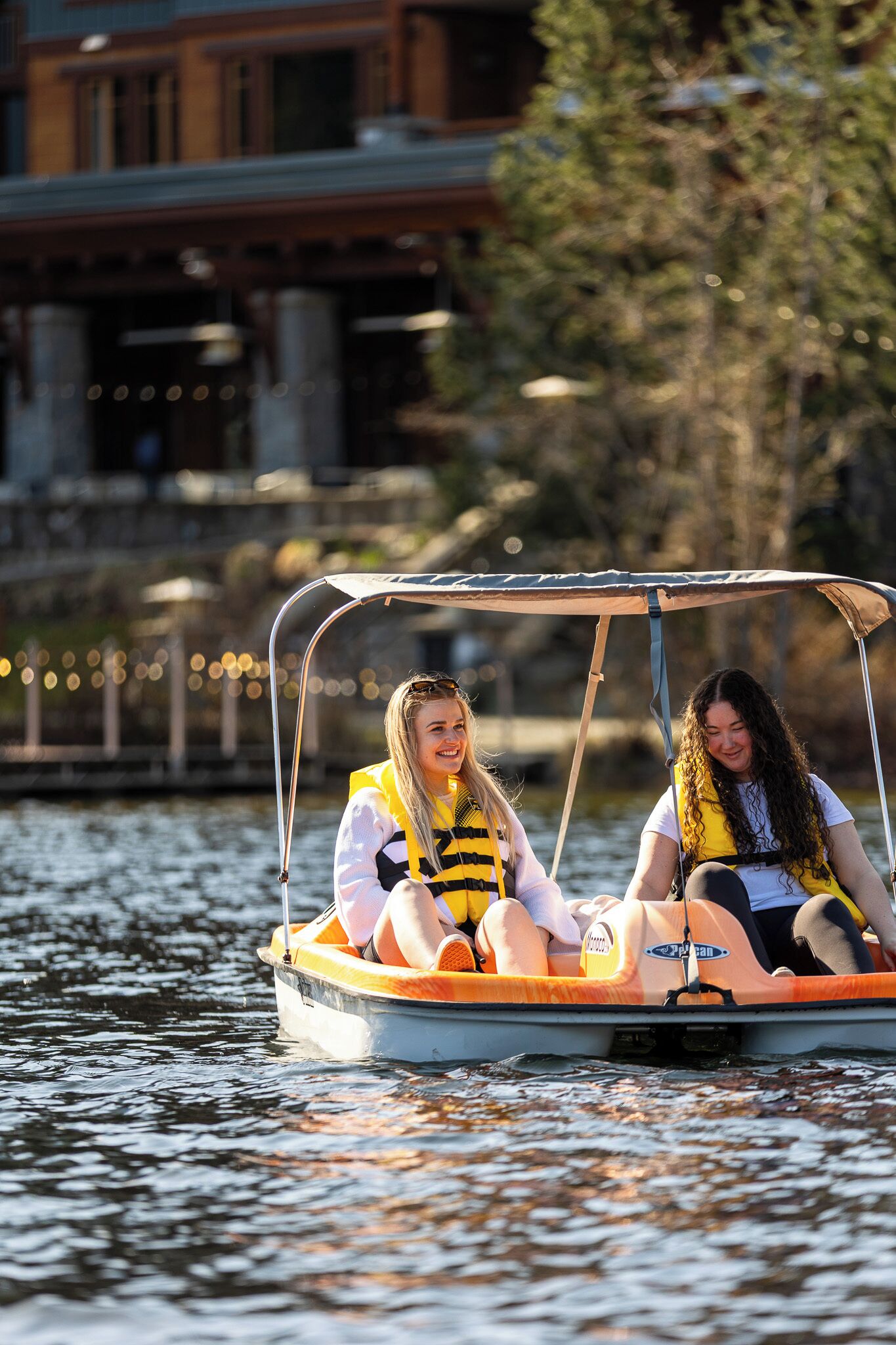 Two people on a boat with a canopy, wearing life jackets, on a body of water with buildings and trees in the background.