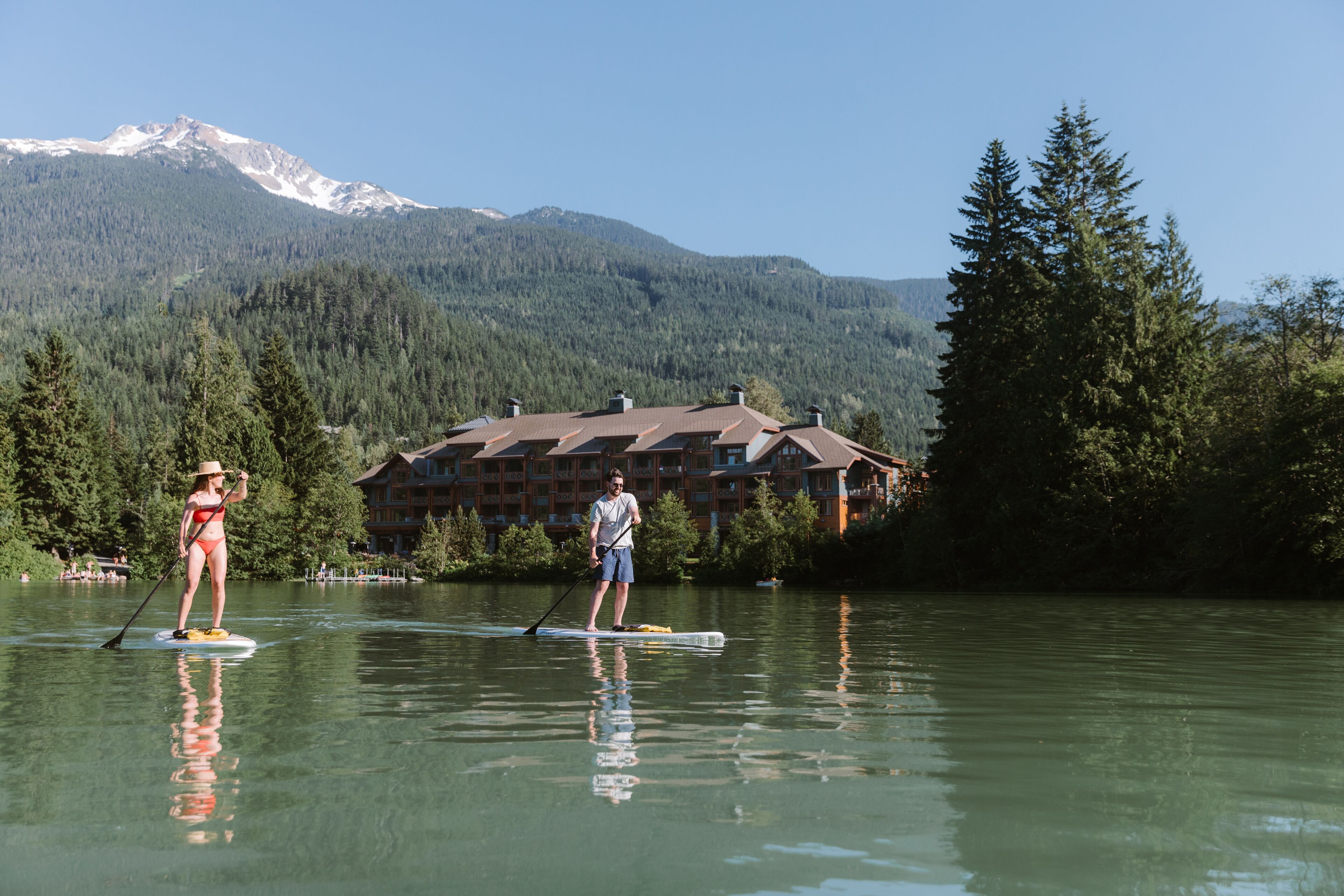 Zwei Personen paddeln auf einem ruhigen See, im Hintergrund ein Berg und ein mehrstöckiges Holzhaus.
