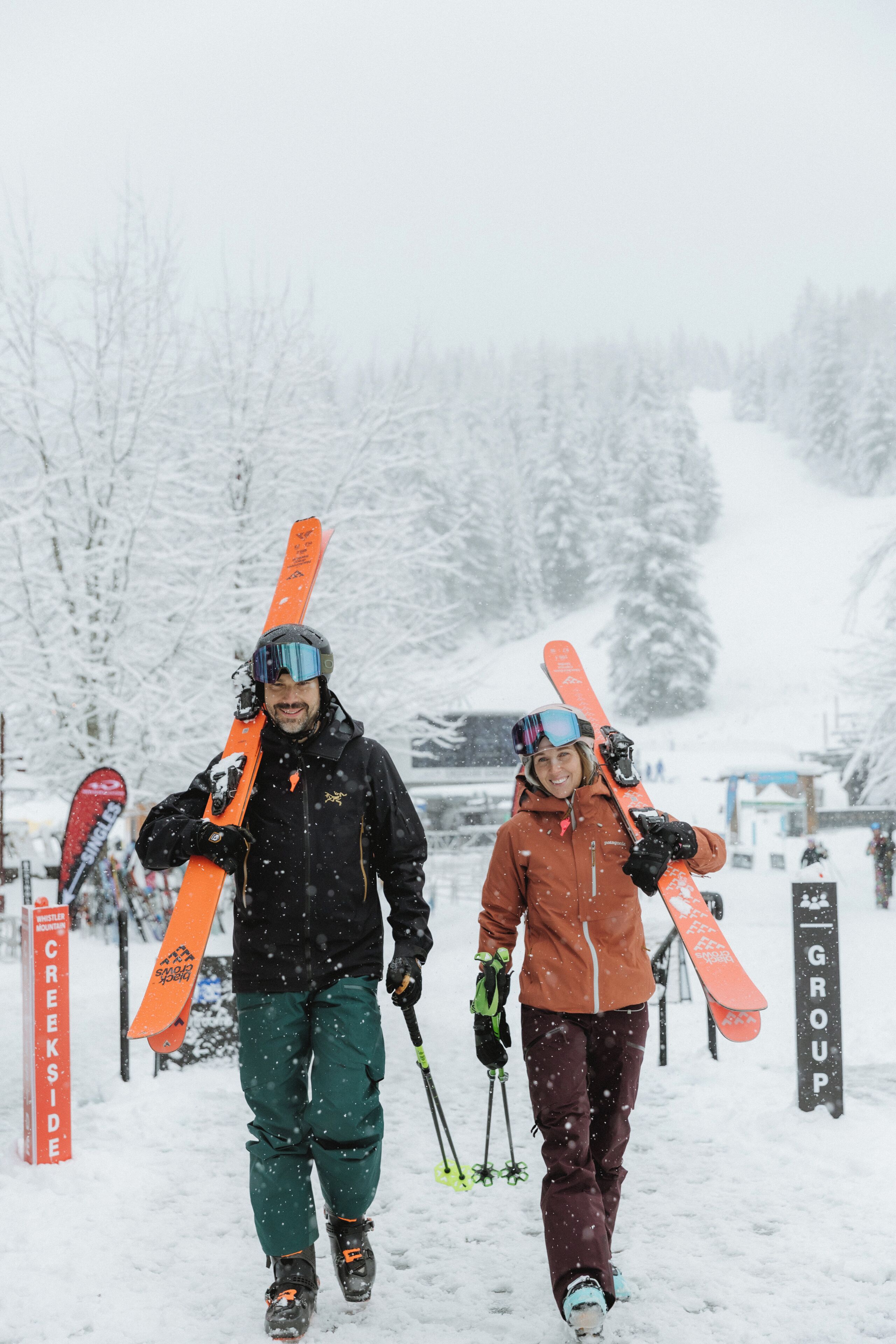 Two people walking with skis, snowboarding gear, and poles.
