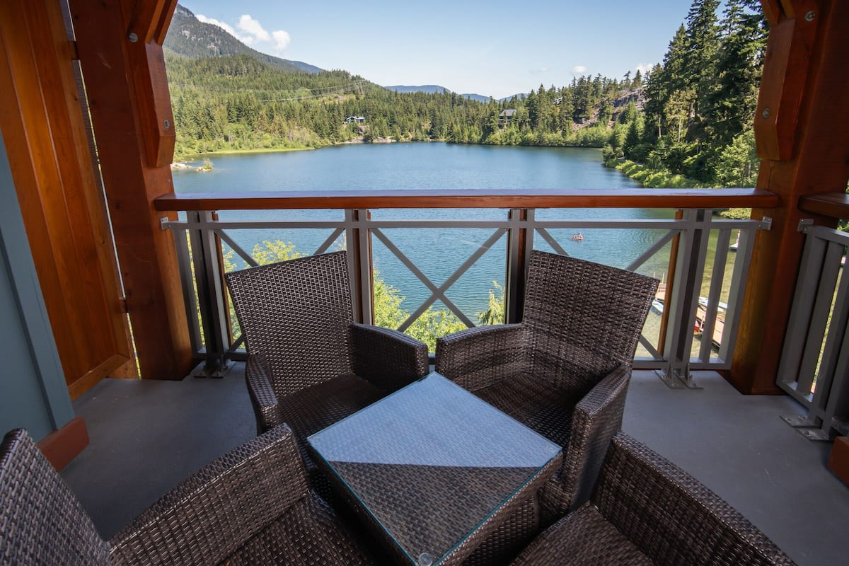 Een balkon met rieten meubilair met uitzicht op een meer en beboste bergen.