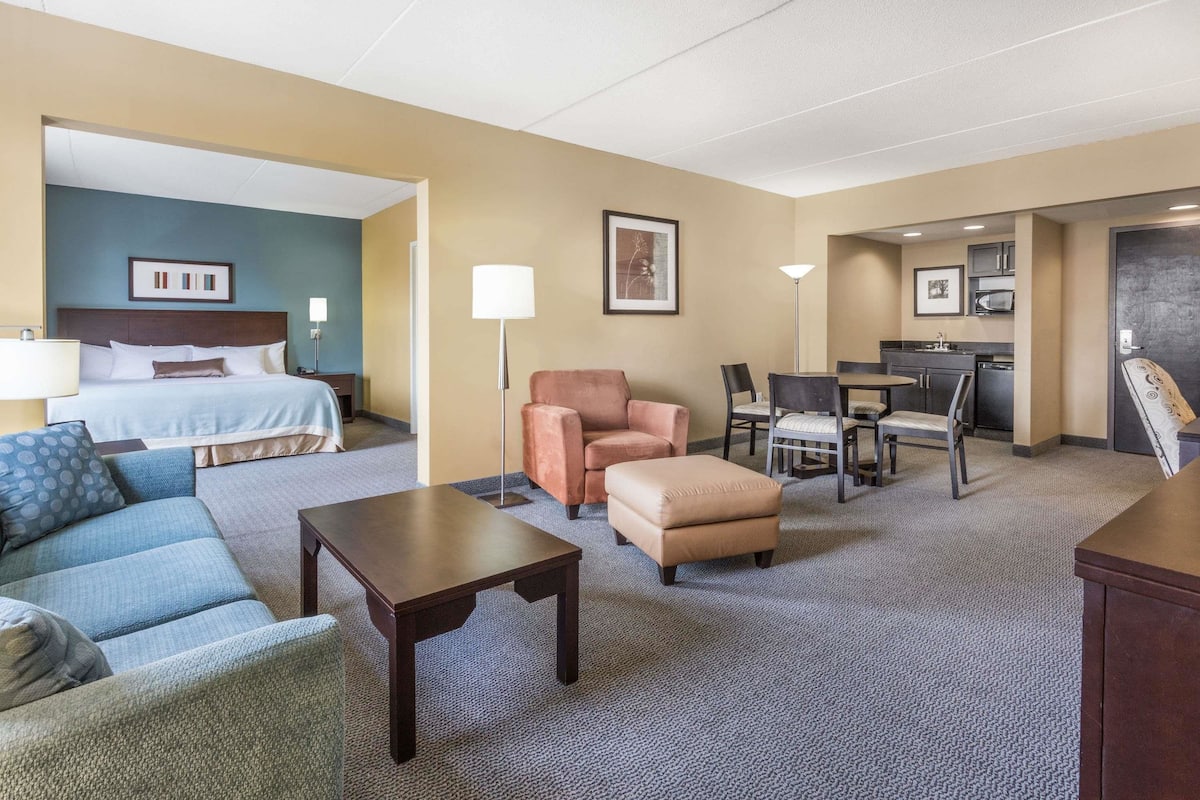 A hotel room with a bed, a blue sofa, a brown armchair, a small table, a lamp, a kitchenette, and a dining area.