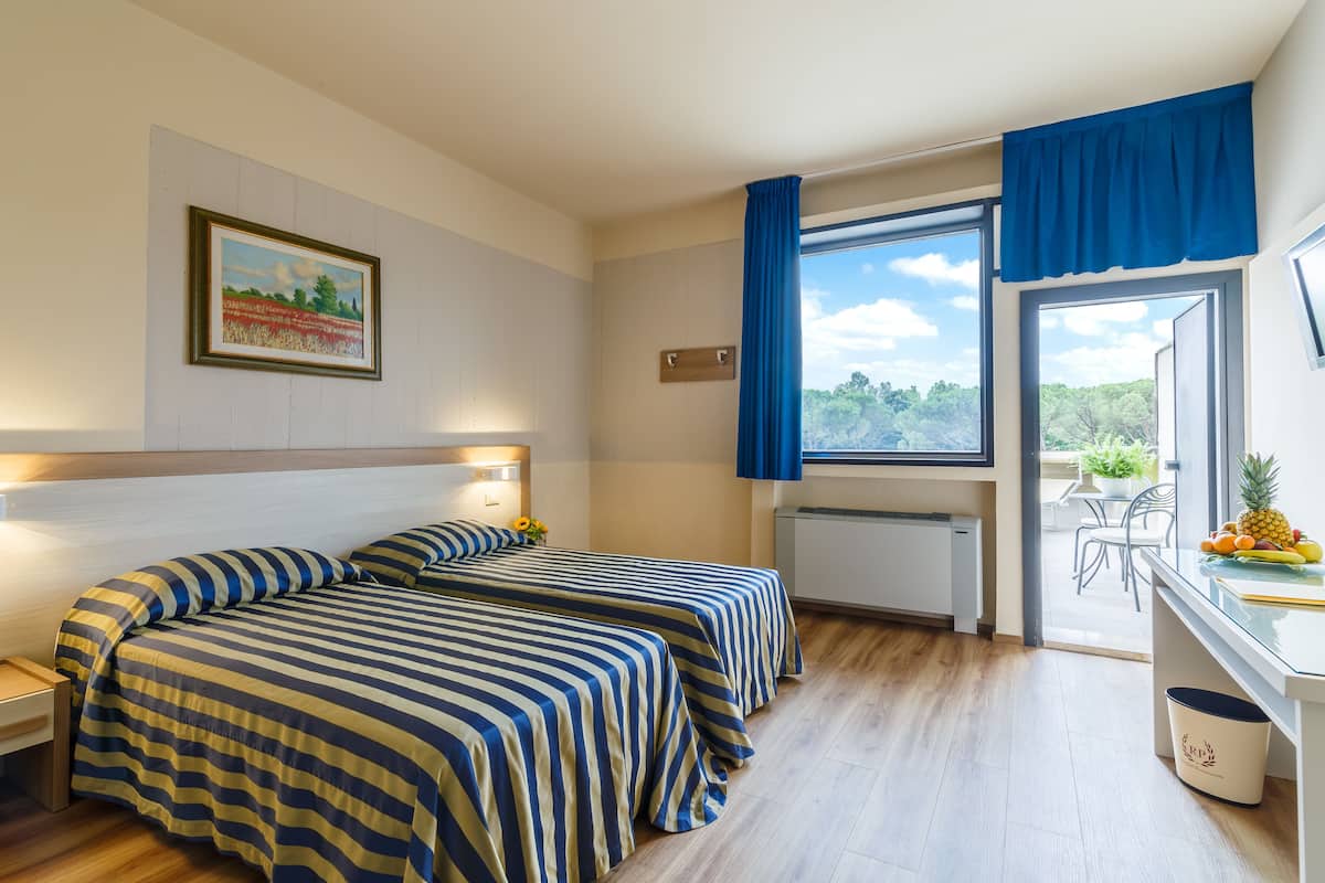 Habitación de hotel con dos camas, un escritorio con fruta, una ventana con vistas y un cuadro en la pared.