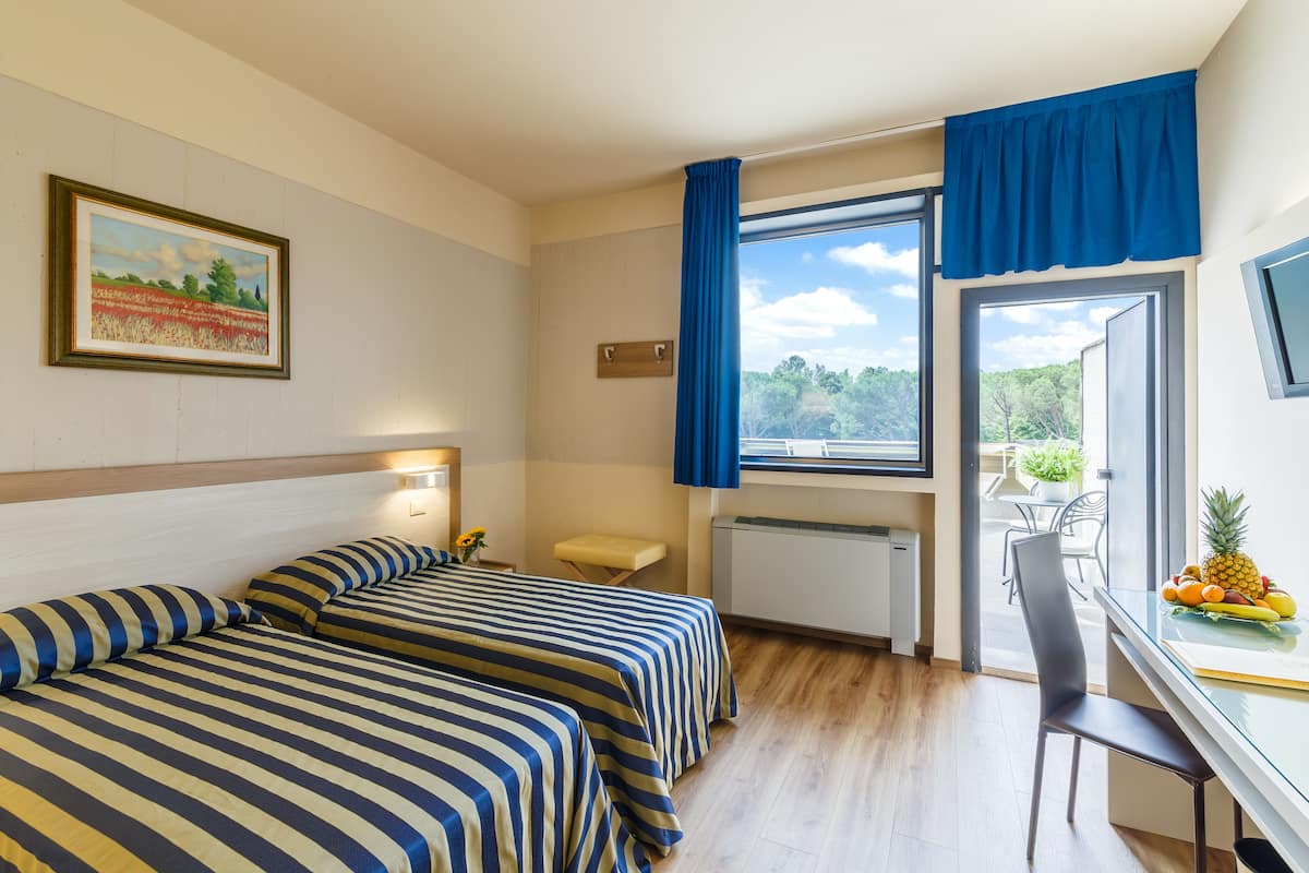 Habitación de hotel con dos camas, un escritorio, una silla y una ventana que da a zonas verdes.