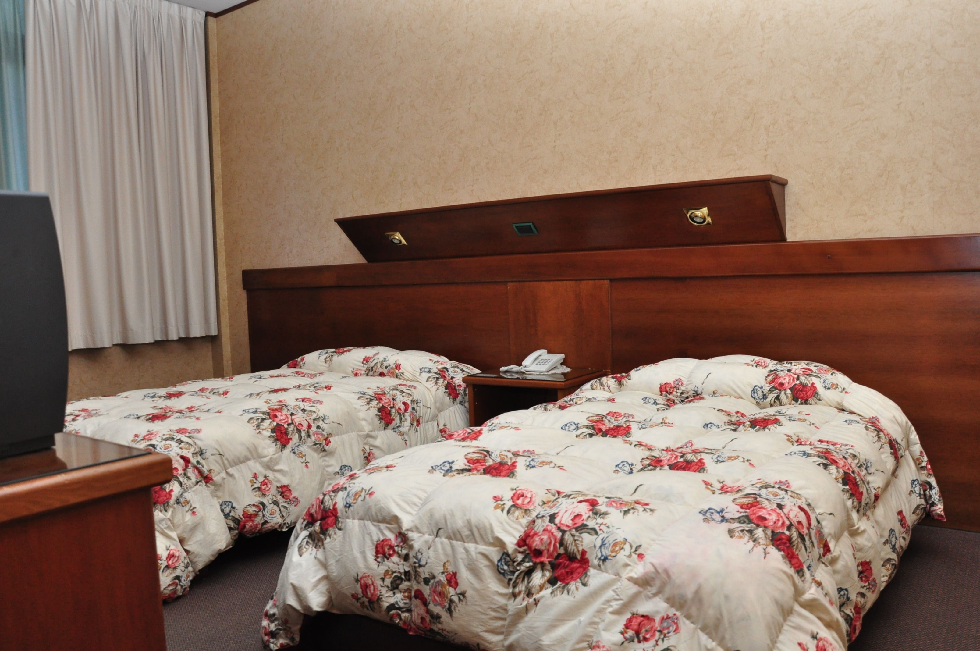 A hotel room with two beds, each with floral bedding, a wooden headboard, a bedside table with a phone, and a television on a desk.