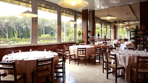 A restaurant with round tables, wooden chairs, and large windows offering a view of greenery. - Hotel Country Club (Capannori)