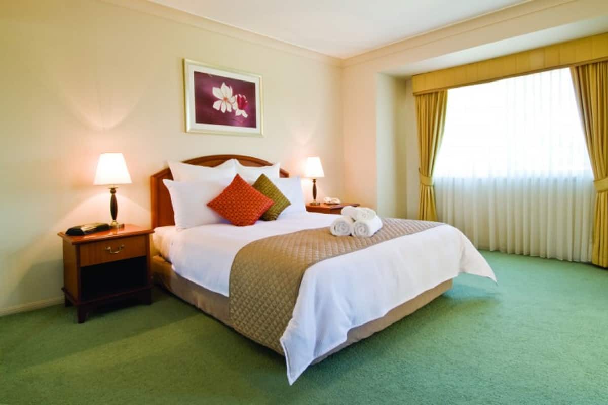 A hotel room with a neatly made bed, two bedside lamps, a framed floral artwork, and a large window with curtains.