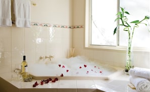 A bathroom with a bathtub filled with water and rose petals, a bottle of wine, and a plant by the window. - Royal Woods Resort (Ashmore)