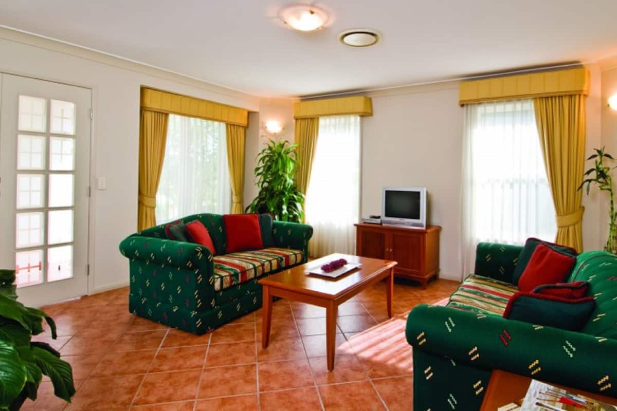 A living room with two green sofas, a wooden coffee table, a television on a wooden cabinet, and large windows with yellow curtains.