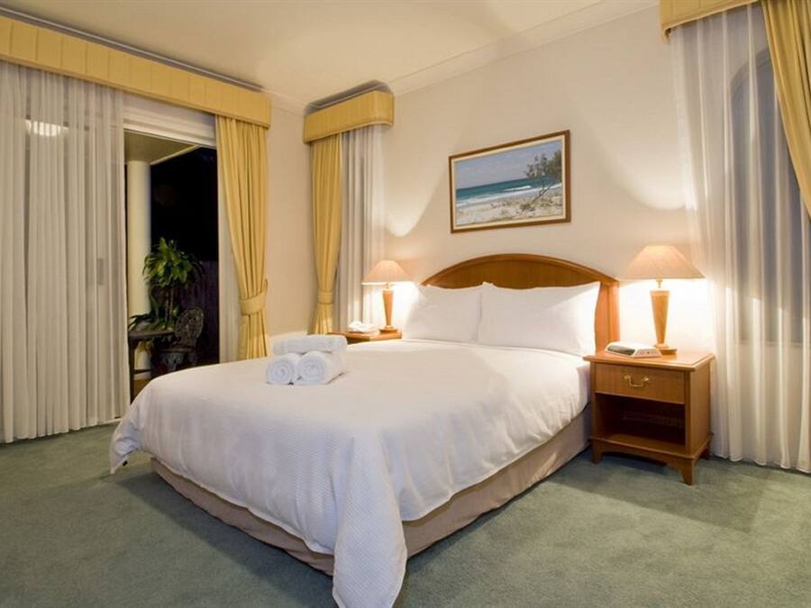 Habitación de hotel con una cama grande, dos lámparas de noche, un cuadro de playa y una puerta que da a otra habitación.