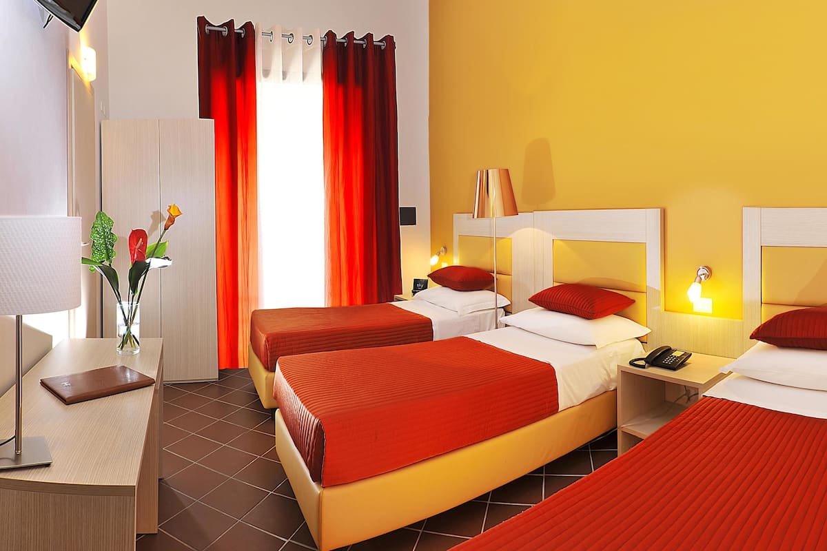 A hotel room with two beds, a desk, a TV, and a window with red curtains.