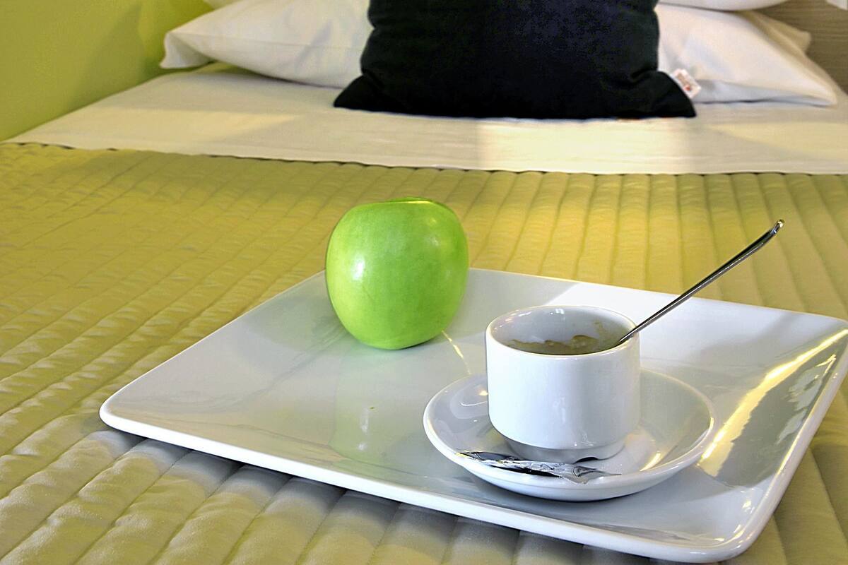 A hotel room with a bed, pillows, a tray with a green apple and a cup of tea, and a painting on the wall.