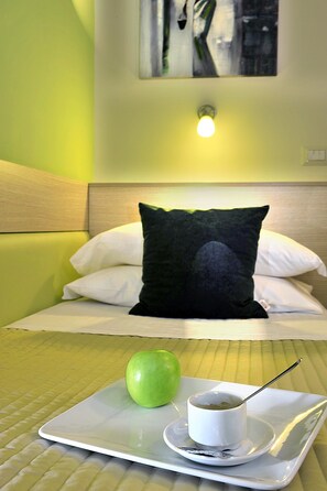 A hotel room with a bed, pillows, a tray with a green apple and a cup of tea, and a painting on the wall.