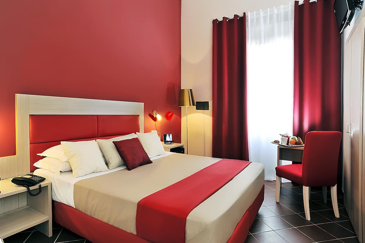 A hotel room with a large bed, a desk with a chair, a nightstand, and a lamp.