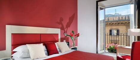 Quarto de hotel com cama vermelha, almofadas brancas, mesa de cabeceira com telefone, cadeira vermelha e vista de um edifício através de uma porta de varanda.