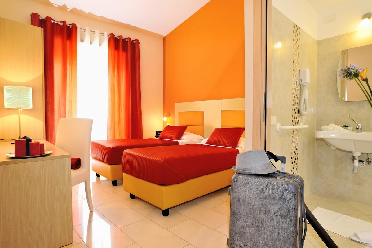 A hotel room with two beds, a desk, a chair, a suitcase, and a bathroom with a sink and mirror.