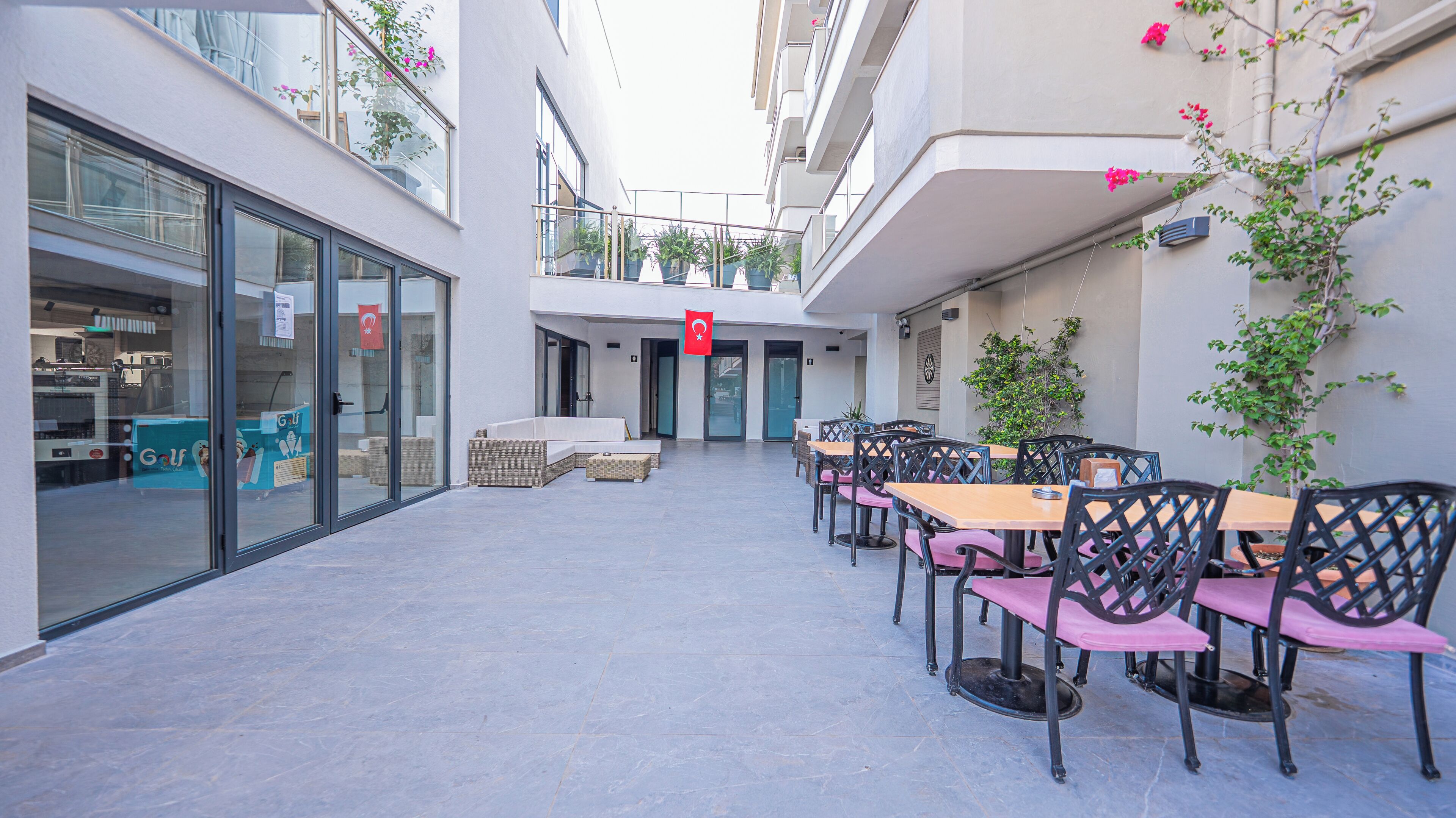 Un área de café al aire libre con mesas y sillas, un escaparate de vidrio a la izquierda y una bandera sobre la entrada.