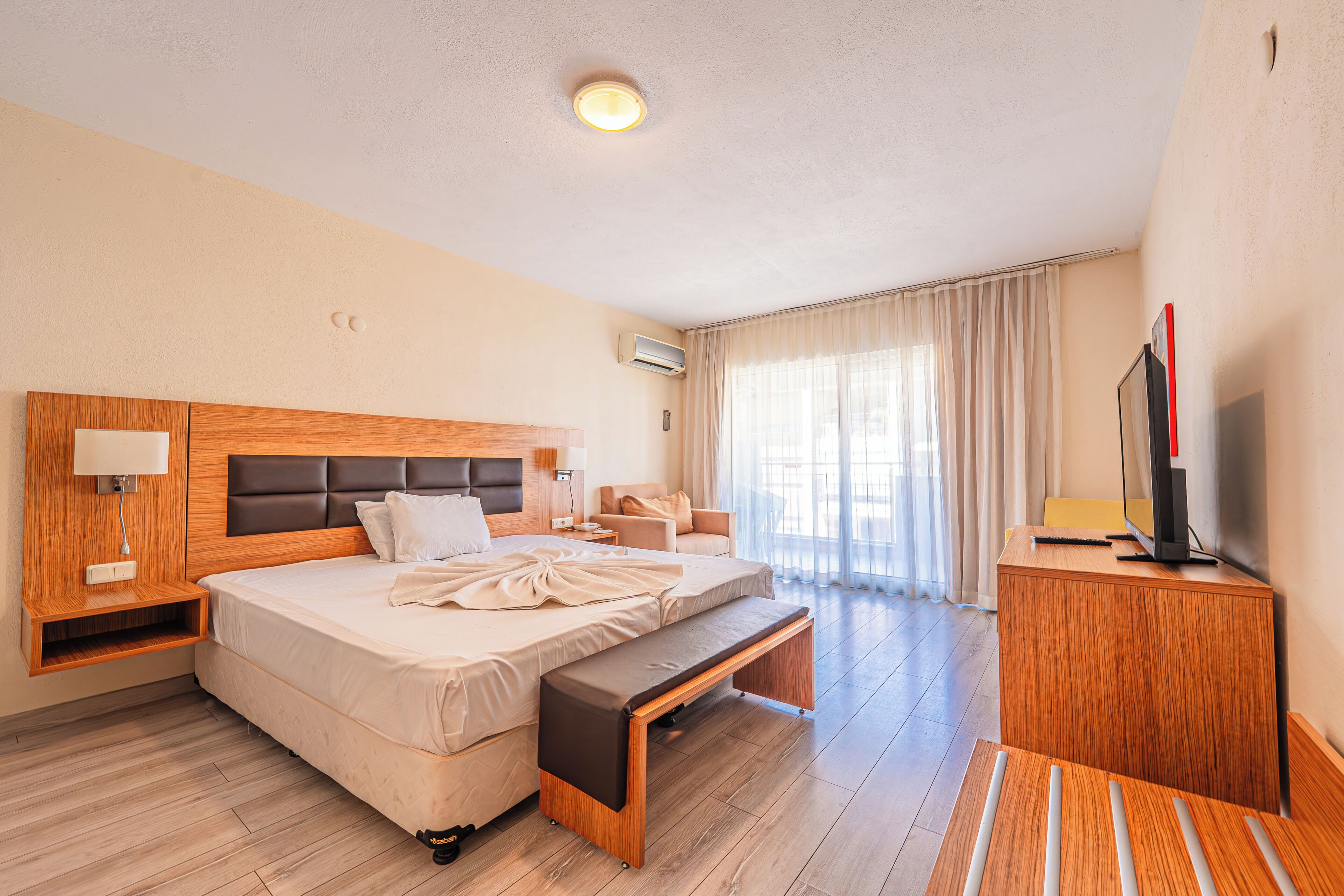 Quarto de hotel com cama, mesa de cabeceira, luminária, ar condicionado e televisão sobre um móvel de madeira.