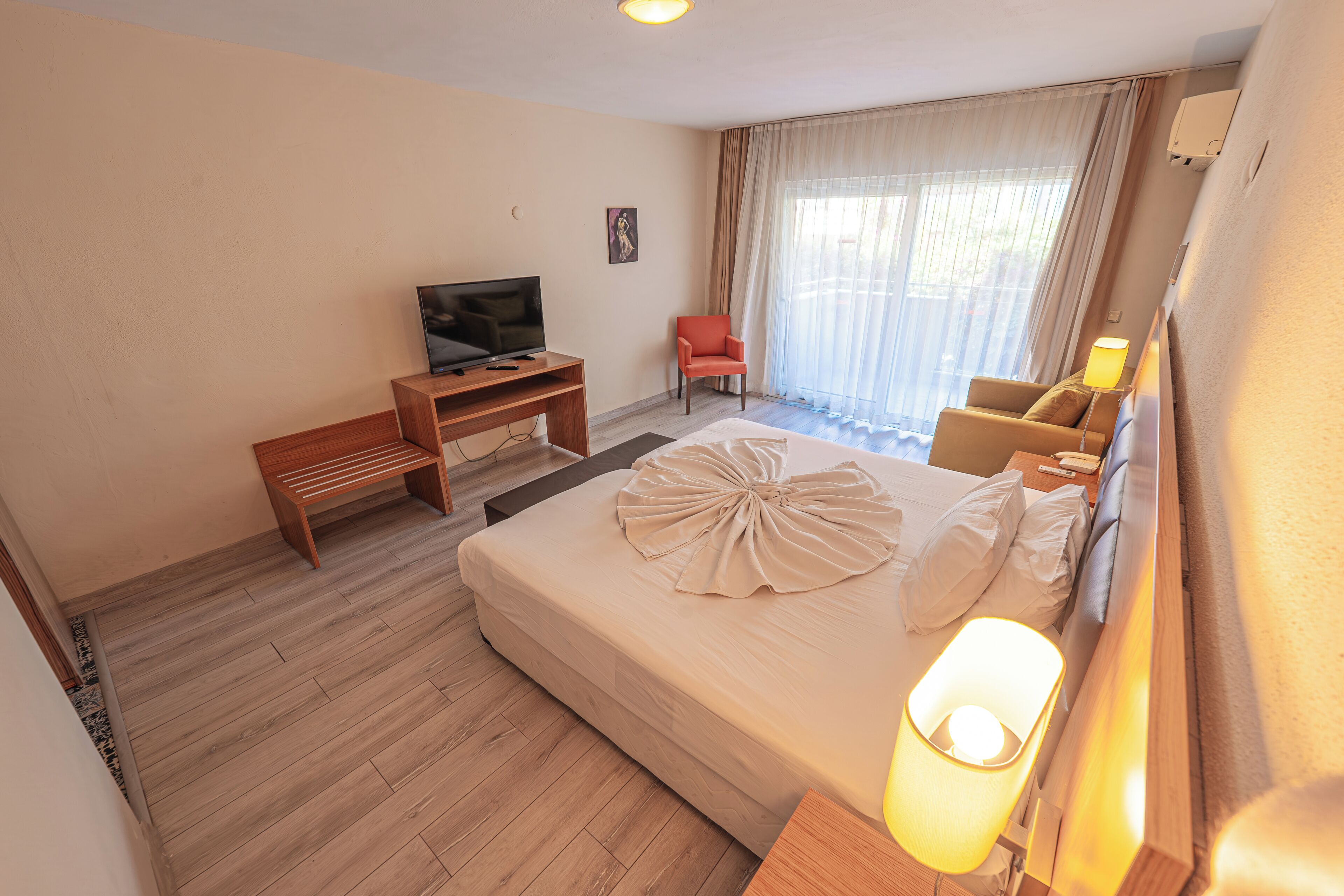 A hotel room with a bed, a TV on a wooden stand, a chair, a desk, and a window with curtains.