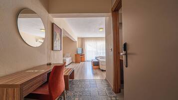 A hotel room with a wooden desk, a red chair, a large mirror, and a view of the living area.