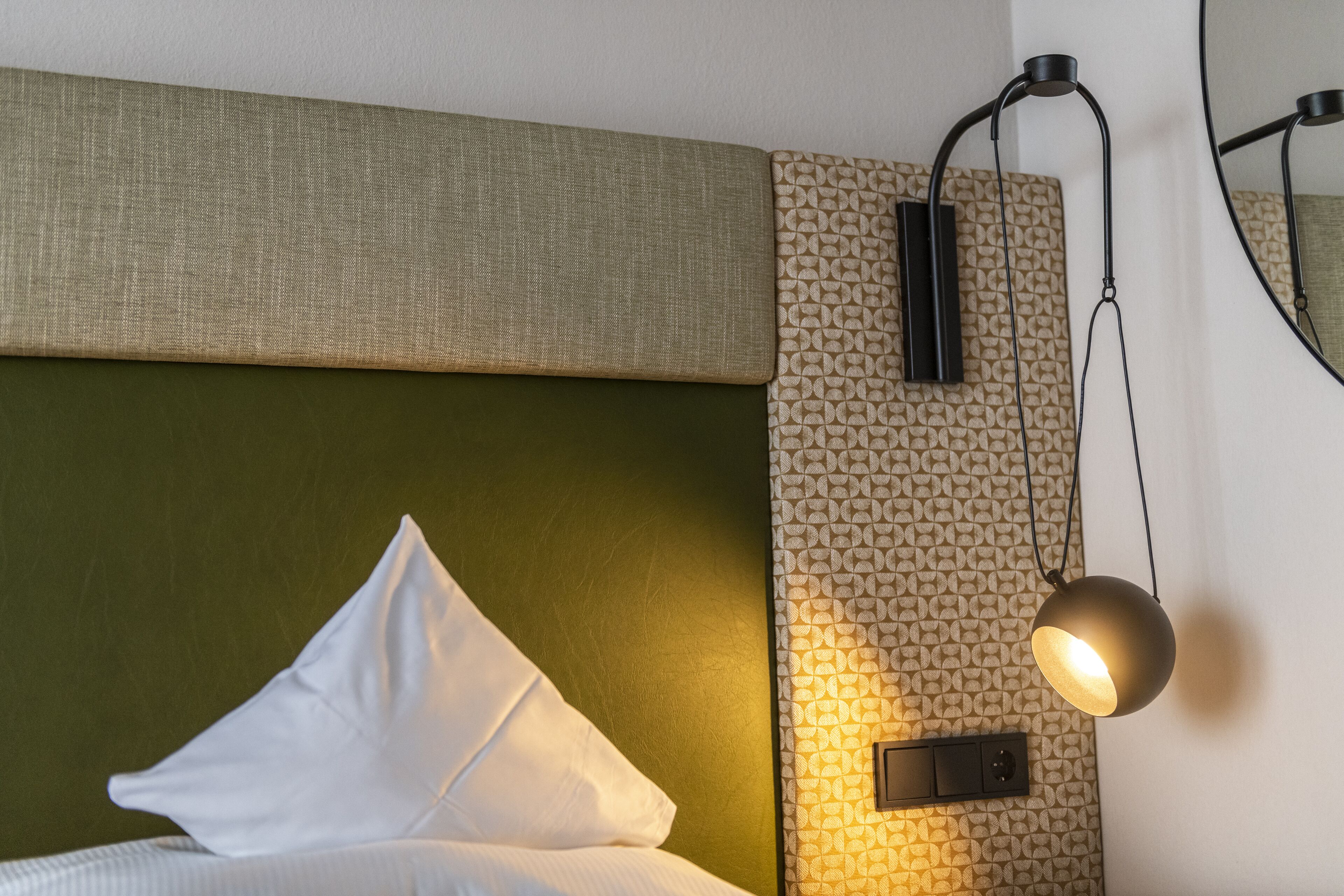Un lit bien fait, avec des oreillers blancs et une tête de lit verte, un mur orné d’un motif et une suspension lumineuse.