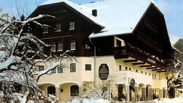 A traditional wooden building with a snow-covered roof, a sign indicating it's a hotel, and a mountainous backdrop. - Rennrad- & Triathlonhotel Jakob (Fuschl am See)