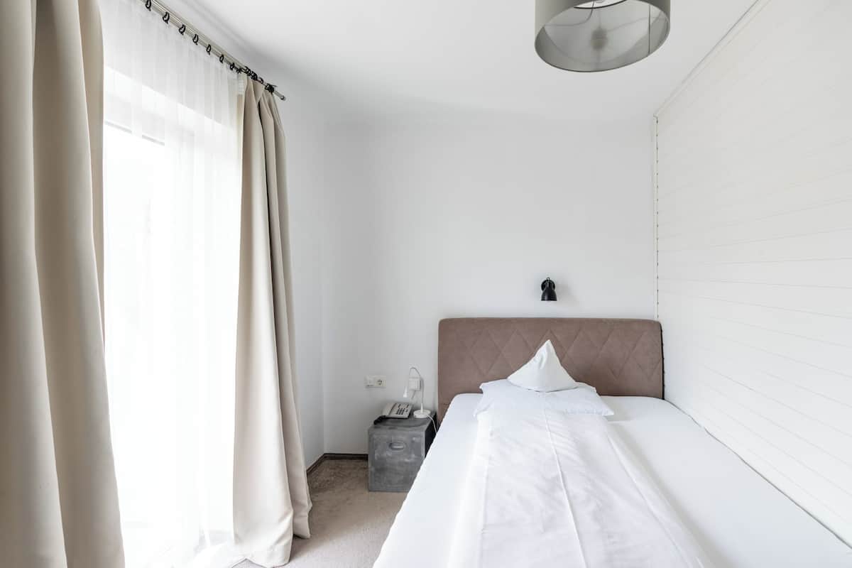 A minimalist hotel room with a single bed, a bedside table with a lamp, and a small nightstand with a phone.