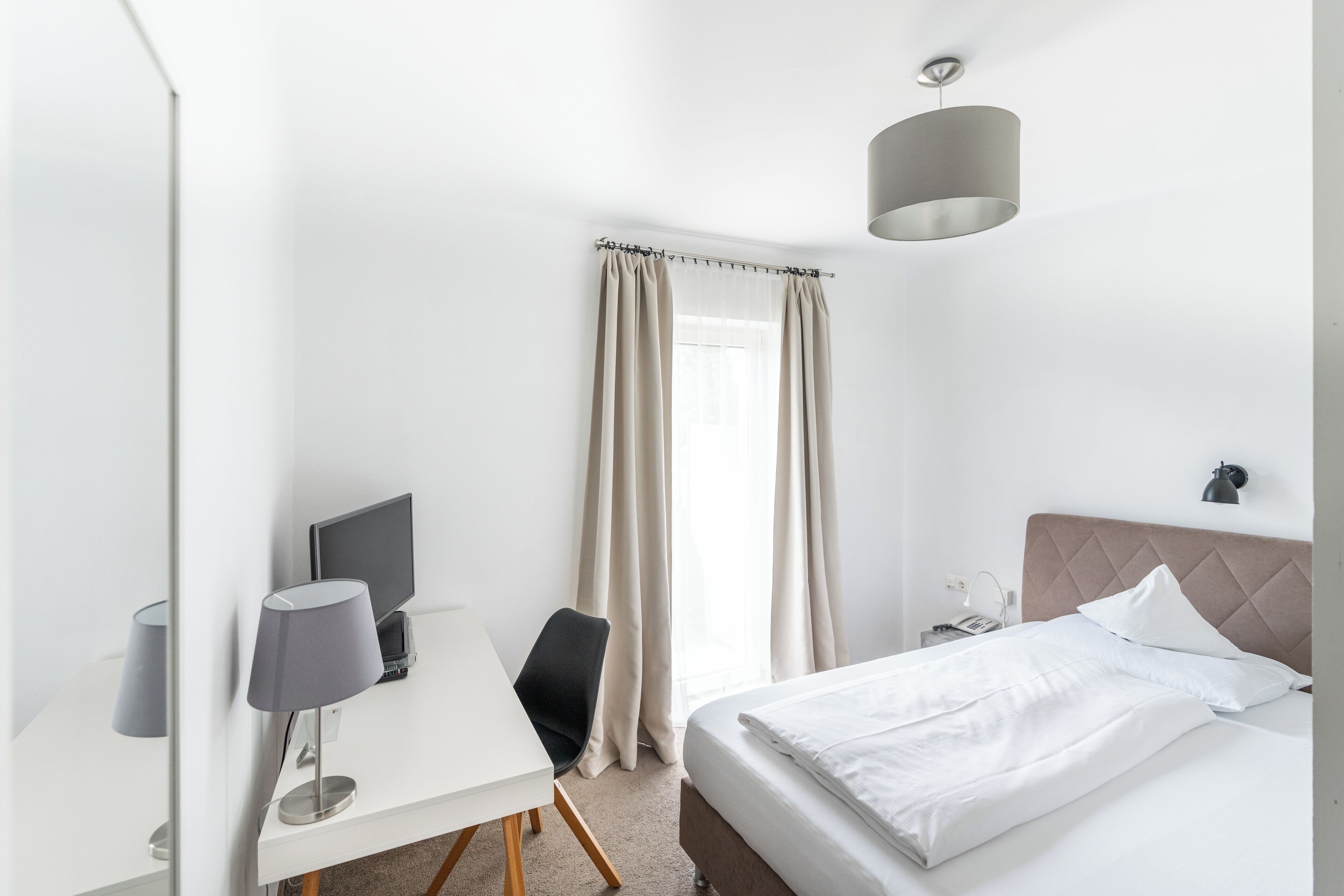 Habitación de hotel con cama, escritorio con lámpara, una silla, ventana con cortinas y lámpara de pared.