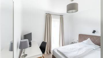 Habitación de hotel con cama, escritorio con lámpara, una silla, ventana con cortinas y lámpara de pared.