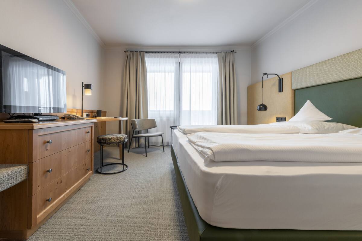 A hotel room with a bed, a desk, a chair, a dresser, a television, and a window with curtains.