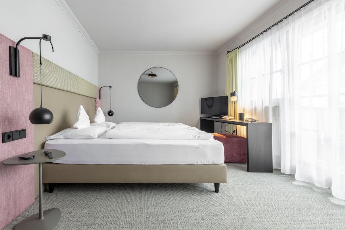 A modern hotel room with a large bed, a round mirror, a desk with a TV, and a view of the outdoors through sheer curtains.