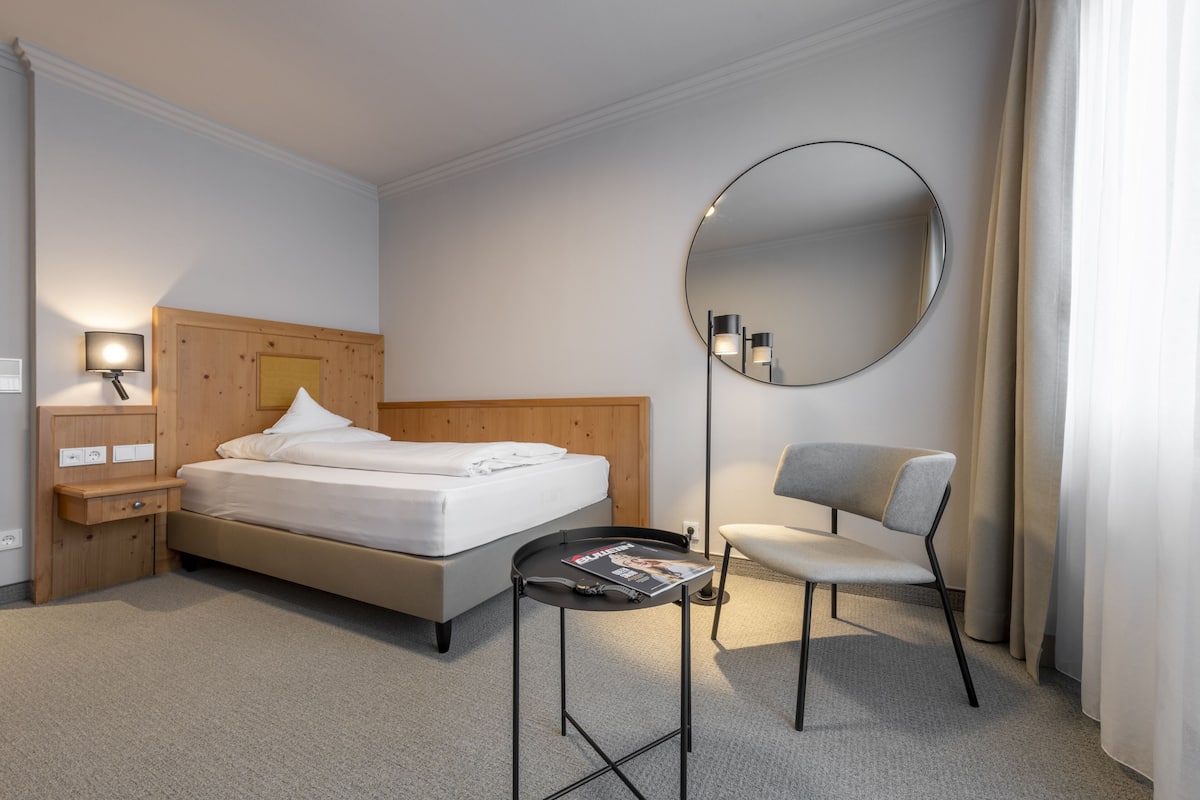 A hotel room with a bed, a bedside table with a lamp, a chair, a round mirror, and a small table with a magazine.