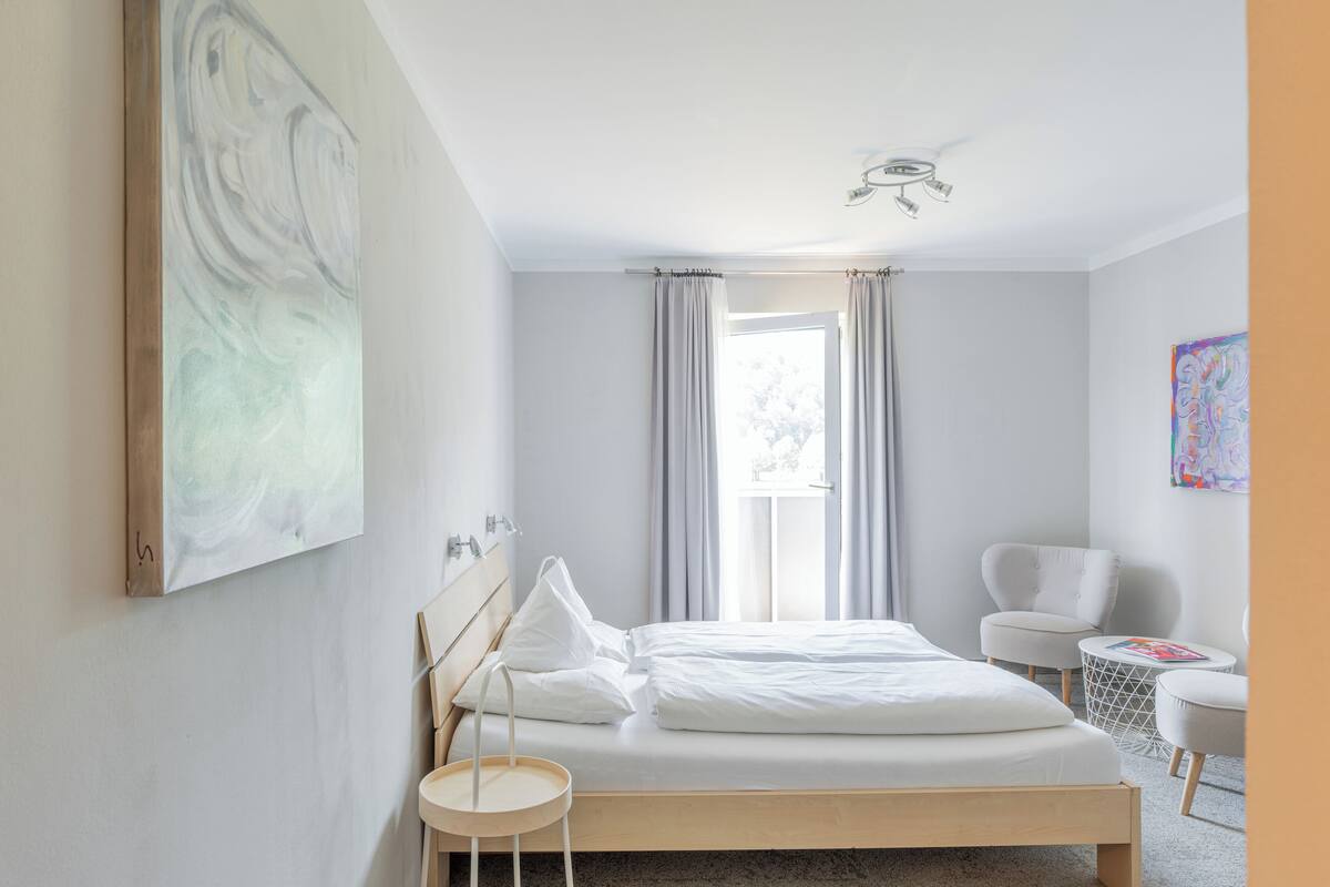 A modern hotel room with a neatly made bed, a small round table, a chair, and a window with curtains.