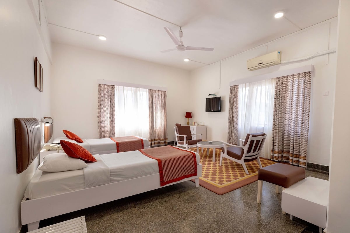 A hotel room with two beds, a desk, a chair, a sofa, a ceiling fan, and an air conditioning unit.