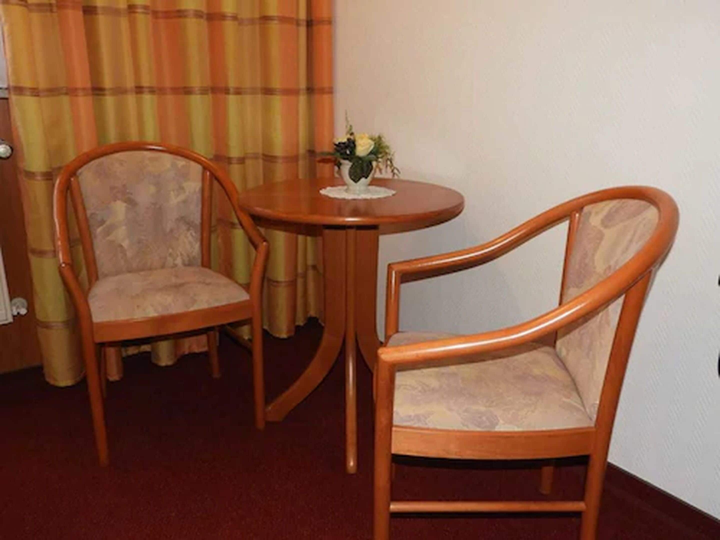 Zwei Holzstühle mit gepolsterten Sitzen und Rücklehnen, ein kleiner runder Tisch mit Blumenvase und ein karierter Vorhang im Hintergrund.
