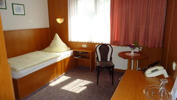 A compact hotel room with a single bed, a small desk, a chair, a table with a vase of flowers, and a telephone.