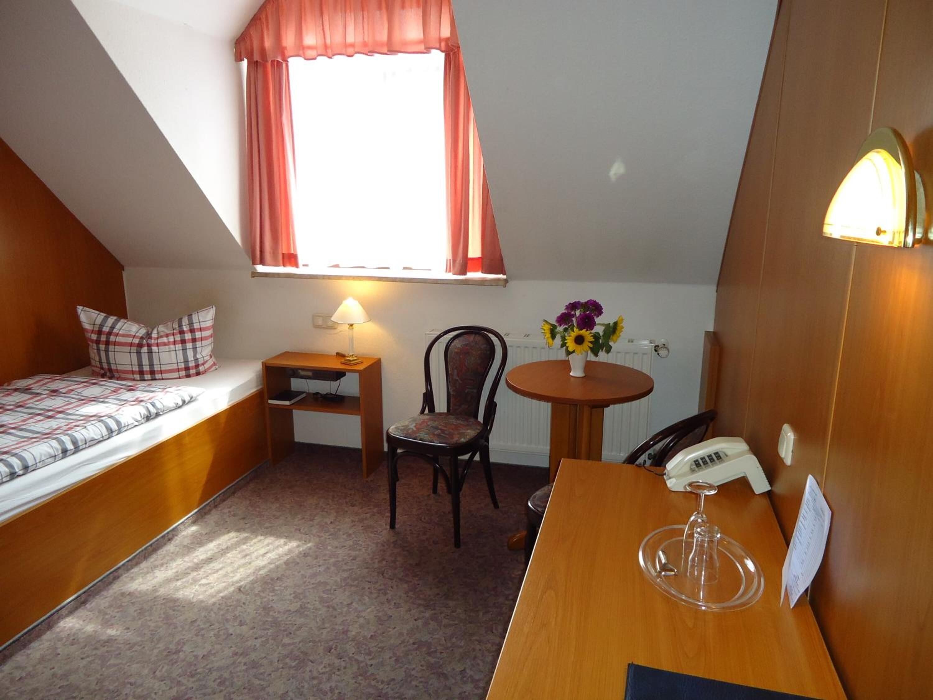 A small, cozy room with a bed, a desk, a chair, a table with a vase of flowers, and a telephone.
