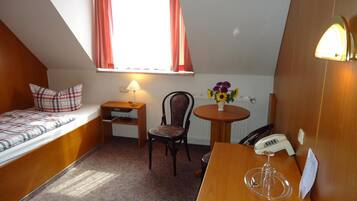 A small, cozy room with a bed, a desk, a chair, a table with a vase of flowers, and a telephone.
