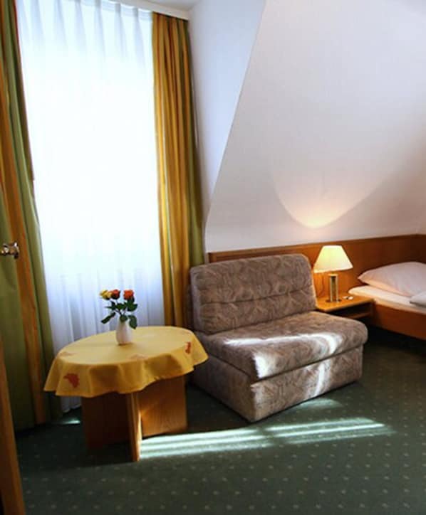 Habitación de hotel con una cama individual, un sillón pequeño, una mesita de noche con lámpara y una ventana con cortinas.