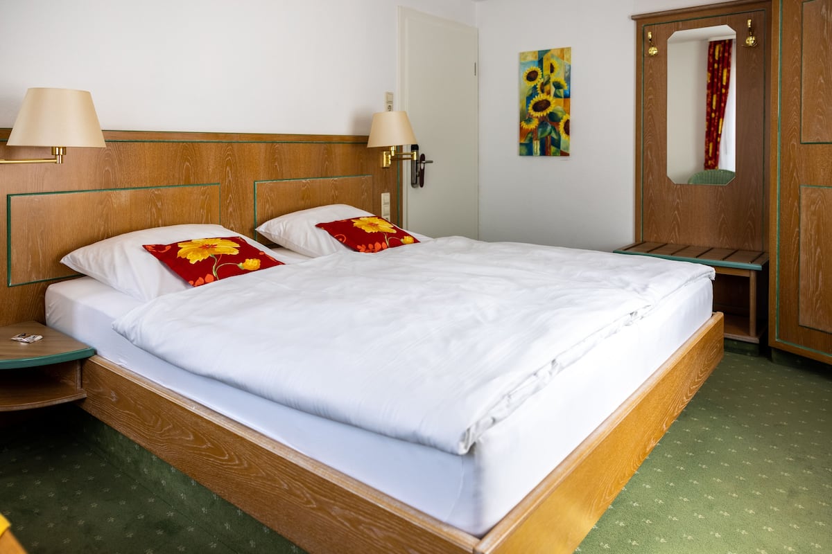 A neatly made bed with white linens and two pillows, one with a red floral design. There are two bedside lamps with beige shades and a wooden headboard. A mirror is visible on the wall to the right.