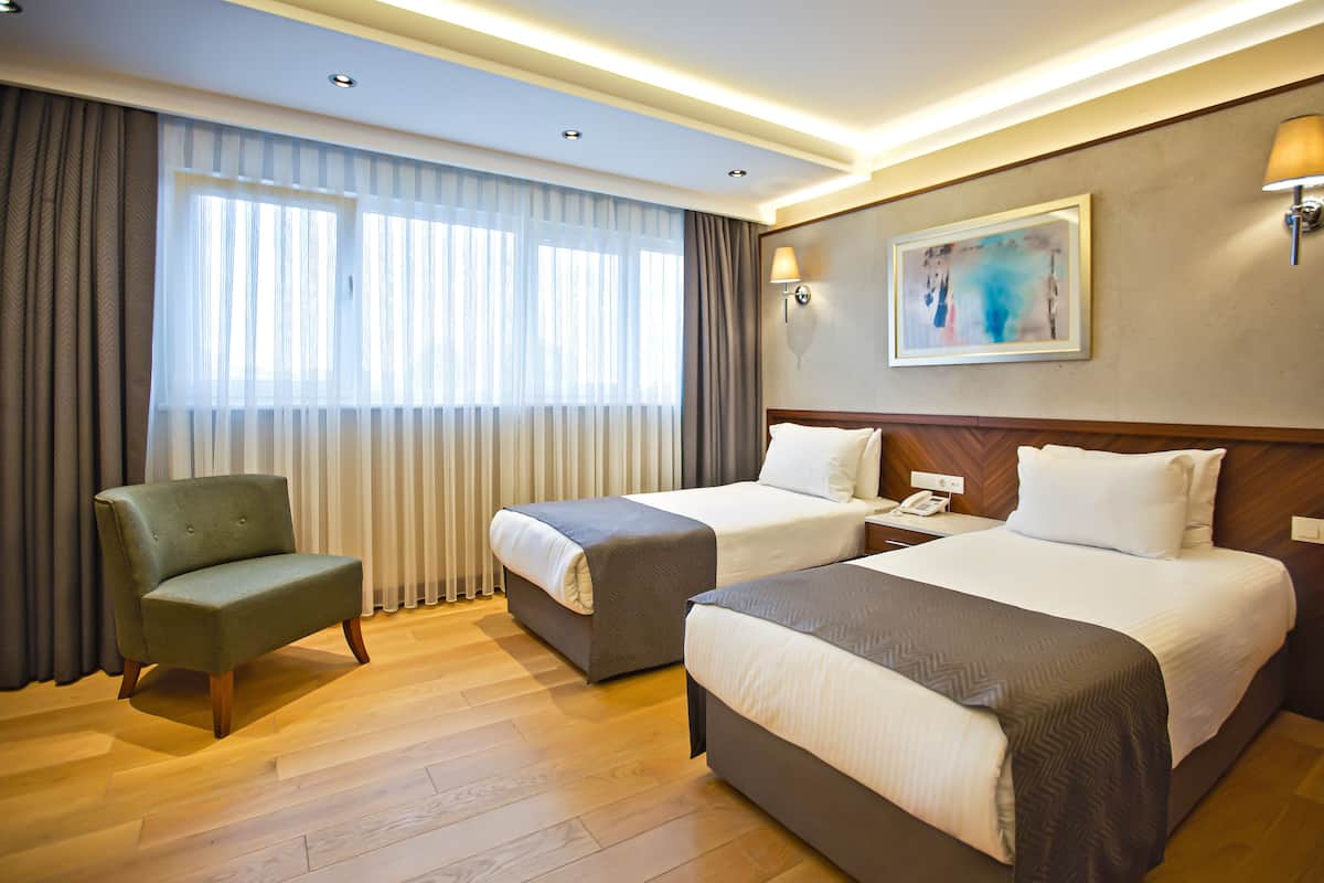 A hotel room with two beds, a green armchair, a painting on the wall, and a window with curtains.